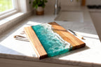 A rectangular wooden cutting board with a turquoise resin wave design. The board has a natural wood grain finish on one side and a simulated ocean wave on the other. A small knife rests on the counter next to the board.