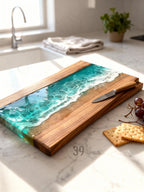 Wooden serving board with a resin ocean design. The board features a rectangular shape with a wooden frame and a turquoise and white resin wave design. A small knife rests on the board, with crackers and grapes nearby. The board is approximately 18 inches long.