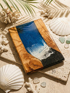 A rectangular wooden serving board with a blue and white resin design resembling ocean waves. The board has a natural wood grain border and a black resin section. Decorative seashells and coral surround the board.