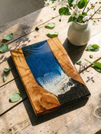 Wooden cutting board with a blue and white resin design resembling ocean waves. The board has a natural wood grain border and the text "The Johnsons EST 2019". A decorative piece for the kitchen.
