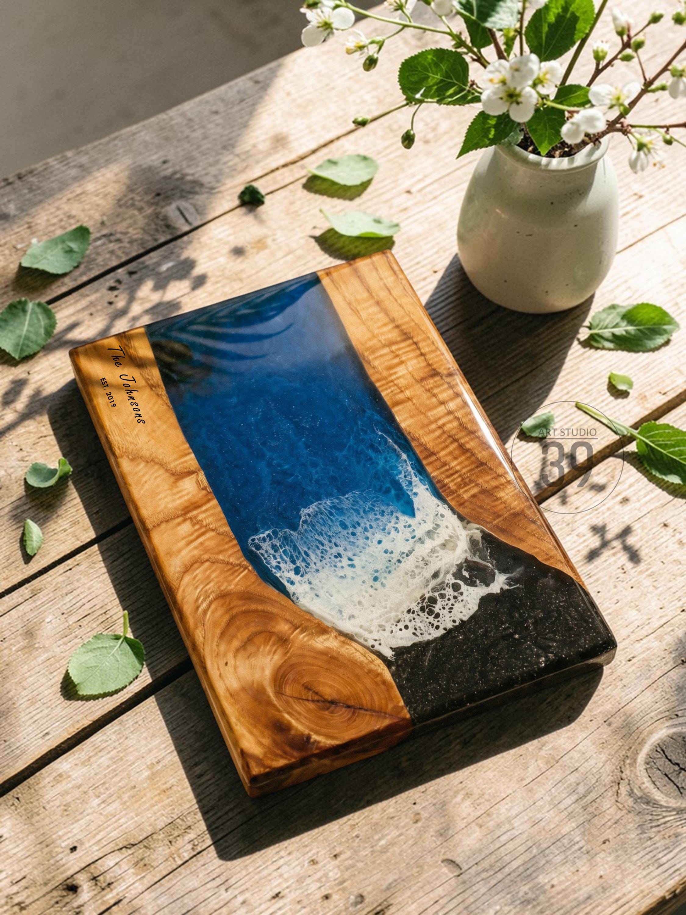 Wooden cutting board with a blue and white resin design resembling ocean waves. The board has a natural wood grain border and the text "The Johnsons EST 2019". A decorative piece for the kitchen.