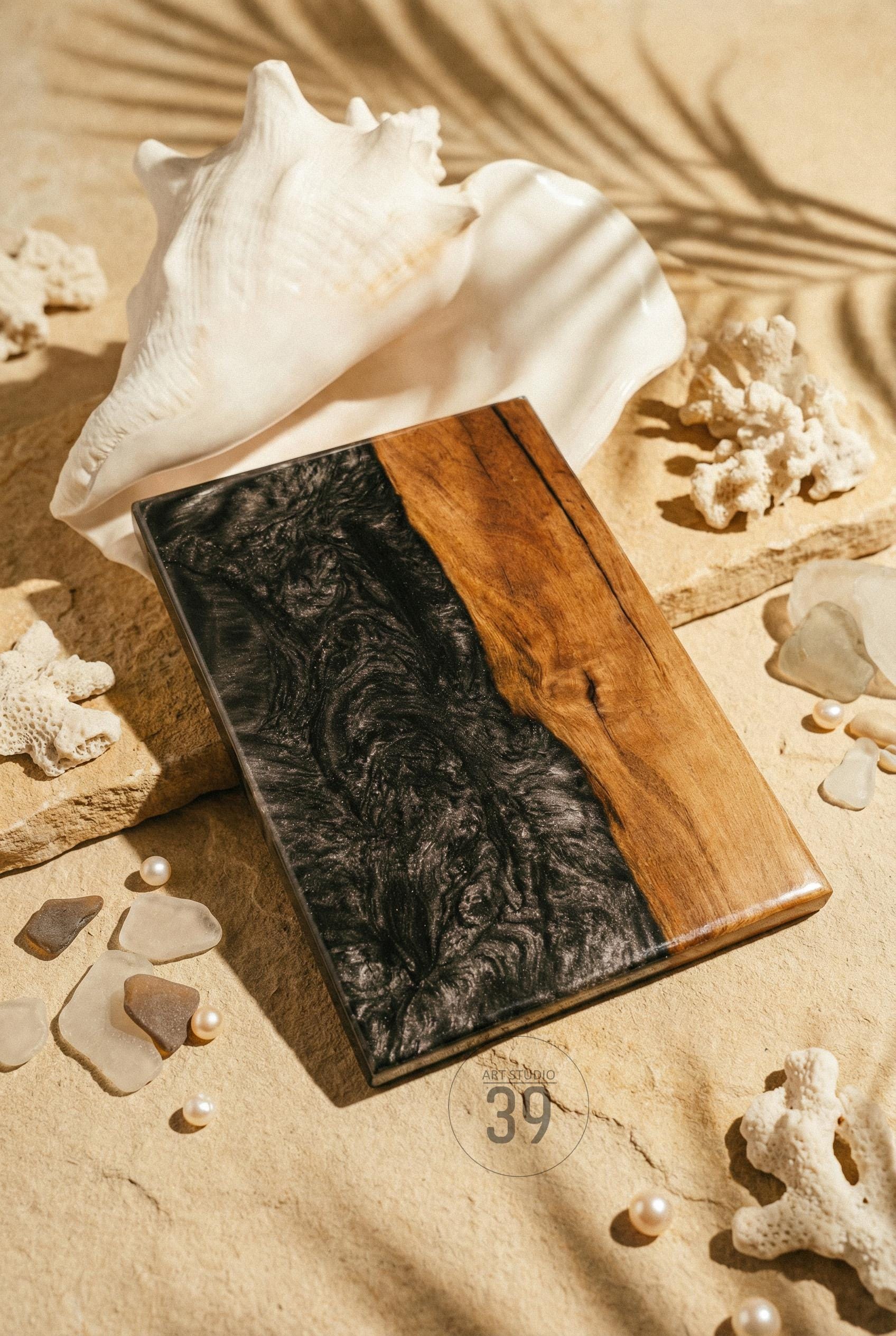 A rectangular wooden serving board with a dark, swirling epoxy resin design on one side and natural wood grain on the other. The board is displayed with seashells, coral, and pearls.