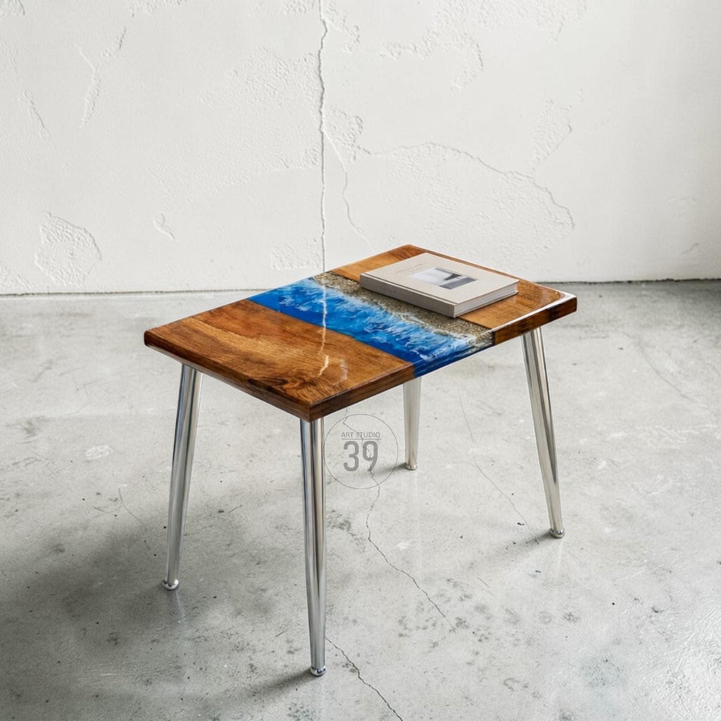 A rectangular side table with a wooden top featuring a blue and white resin design resembling water. The table has four silver metal legs and a book on top. The table is approximately 24 inches long and 16 inches wide.