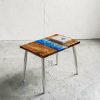 A rectangular side table with a wooden top featuring a blue and white resin design resembling water. The table has four silver metal legs and a book on top. The table is approximately 24 inches long and 16 inches wide.