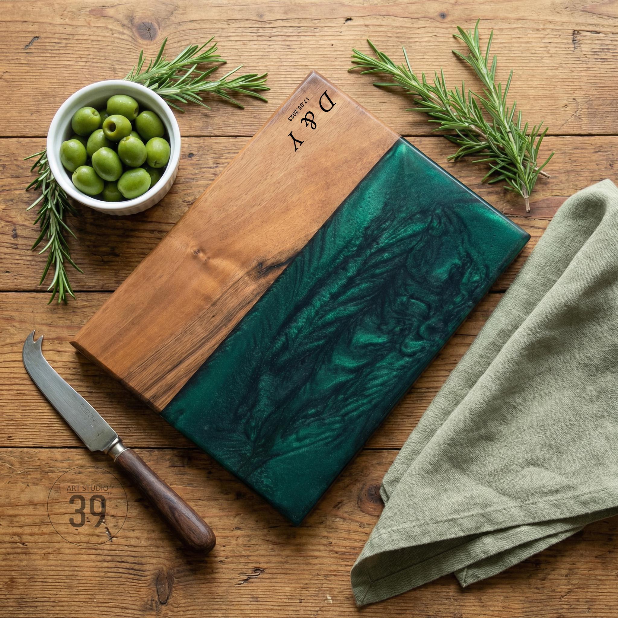 Wooden serving board with a dark green epoxy resin inlay. The board has a natural wood grain finish and is paired with a small cheese knife, a bowl of olives, and sprigs of rosemary. The initials "D &amp; Y" are engraved on the wood.