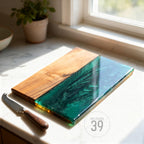 A wooden cutting board with a vibrant green epoxy resin section. A cheese knife with a wooden handle rests beside the board. The board is on a white countertop. The text "ART STUDIO 39" is in a circle on the bottom right.