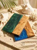 Wooden serving boards with epoxy resin accents in green and blue. The boards are rectangular and feature a natural wood grain. The resin has a swirled, glossy finish. The image also includes seashells and pearls.