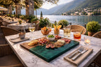 A beautifully set table at an outdoor restaurant overlooking a lake. The table features a charcuterie board with meats, cheeses, and figs, along with drinks and a lit candle. The scene is bathed in warm, golden light.