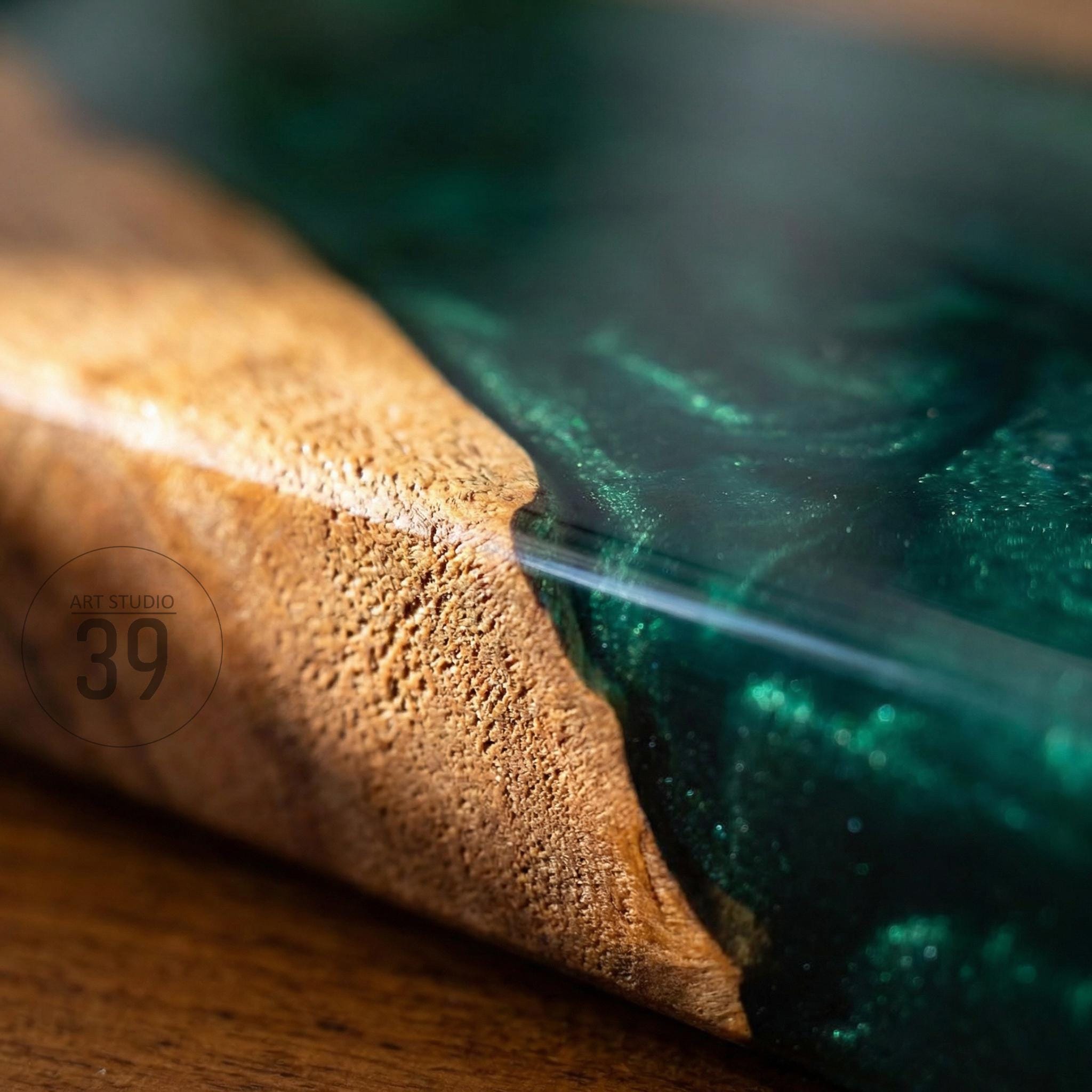 Close-up of a wooden surface with a section of dark green epoxy resin. The wood has a light brown color and a textured grain. The epoxy resin has a glossy finish and swirls of green. The text "ART STUDIO 39" is visible.