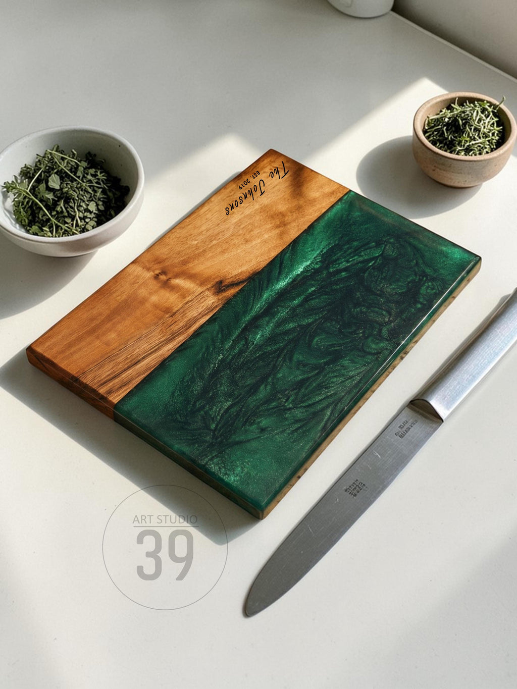 A rectangular wooden cutting board with a section of green epoxy resin. The board has the text "The Johnsons Est 2019" engraved on it. A stainless steel knife and two small bowls with herbs are next to the board.
