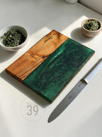 A rectangular wooden cutting board with a section of green epoxy resin. The board has the text "The Johnsons Est 2019" engraved on it. A stainless steel knife and two small bowls with herbs are next to the board.