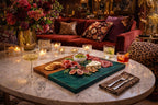 A marble-topped coffee table displays a charcuterie board with olives, cheese, meats, and figs. Champagne and cocktails sit nearby, with candles and a floral arrangement in the background. A red velvet sofa and leopard print accents complete the scene.