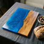 A rectangular wooden serving board with a vibrant blue epoxy resin section. The wood has a natural grain and a warm tone, contrasting with the glossy blue. A small bowl of olives and a loaf of bread are nearby.
