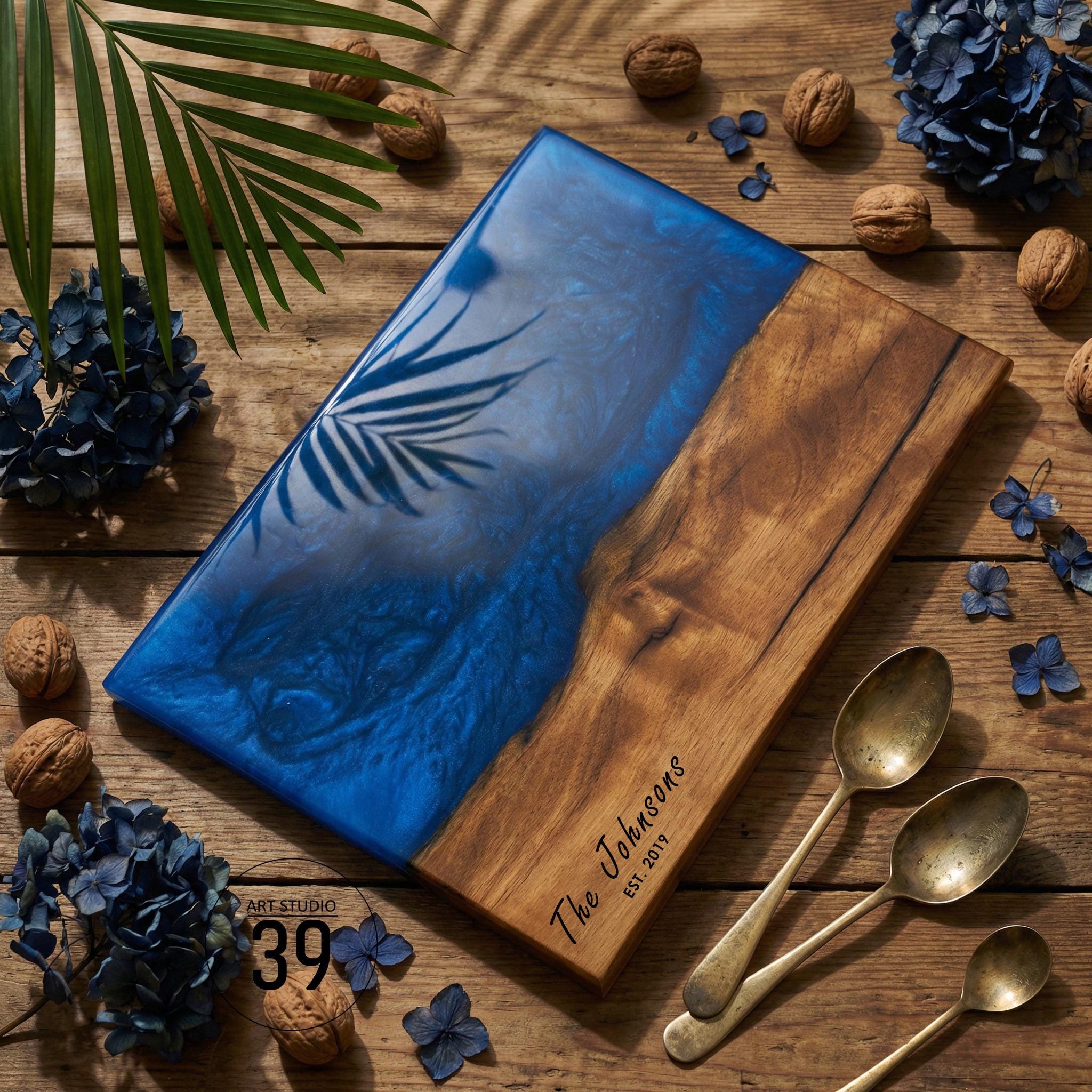 A rectangular wooden serving board with a blue resin section and a palm leaf design. The wooden section is engraved with "The Johnsons EST. 2019". The board is surrounded by walnuts, blue flowers, and vintage spoons.