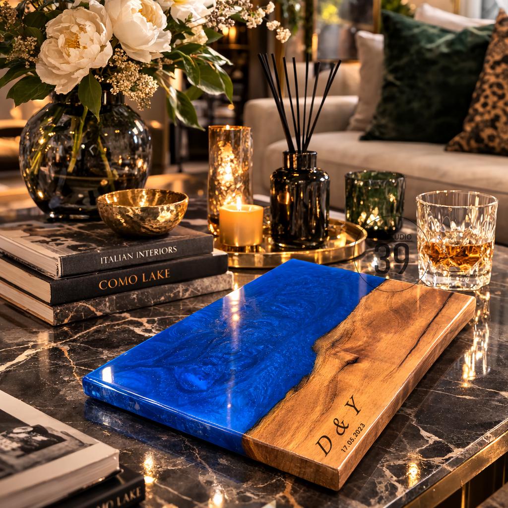 A rectangular wooden serving board with a vibrant blue epoxy resin section. The wooden portion is engraved with the initials "D &amp; Y" and the date "17.05.2023". The board is displayed on a marble-topped coffee table, surrounded by decorative items.