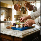 A wooden and blue resin serving board with cheese, grapes, figs, and crackers. A person is garnishing the board with rosemary. The board is on a marble countertop in a kitchen.