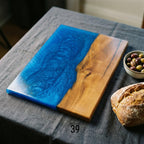 A rectangular wooden serving board with a blue resin section resembling water. The other half is natural wood grain. A small bowl of olives and a loaf of bread are nearby. The board is approximately 12 inches long and 8 inches wide.