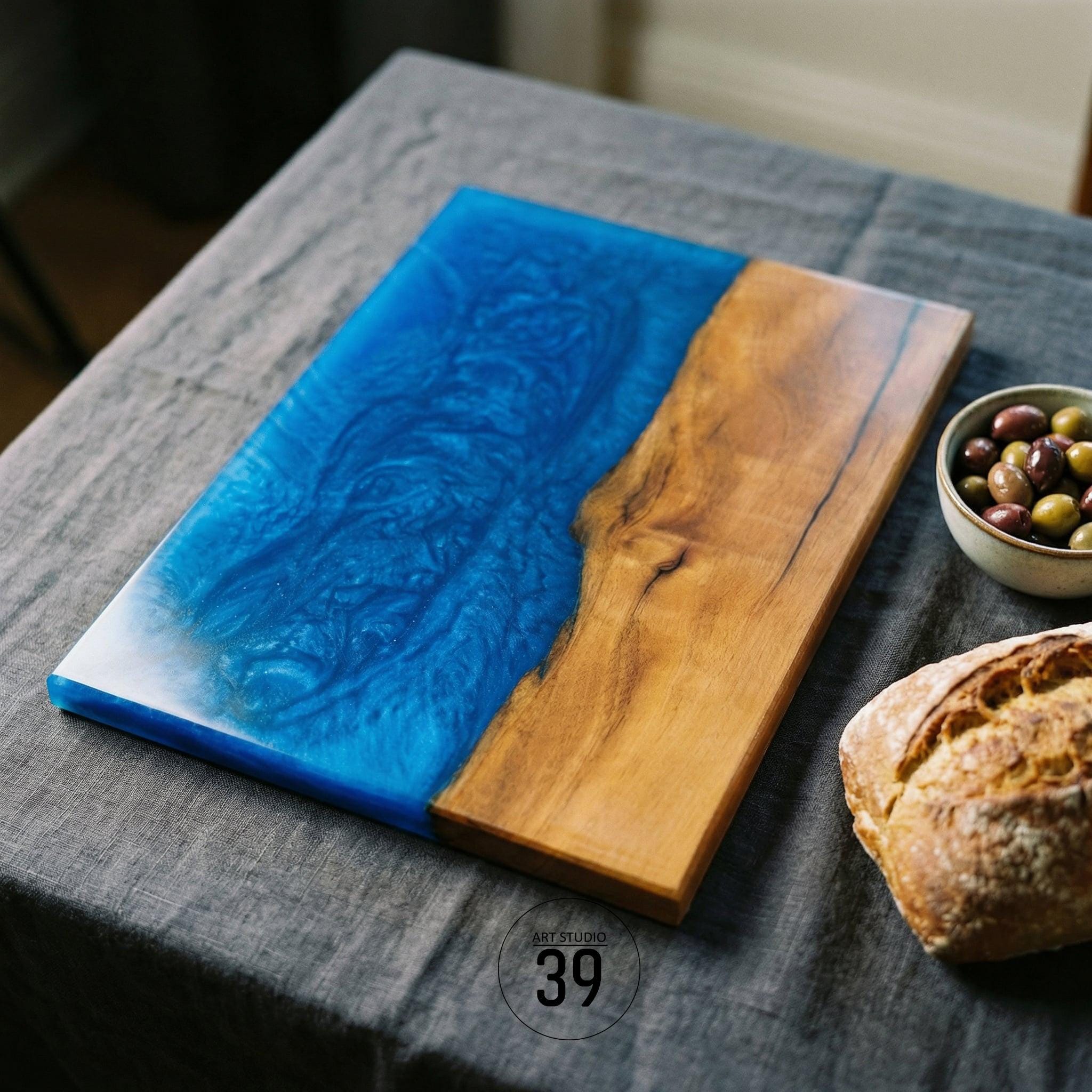 A rectangular wooden serving board with a blue resin section resembling water. The other half is natural wood grain. A small bowl of olives and a loaf of bread are nearby. The board is approximately 12 inches long and 8 inches wide.