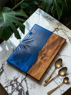 A rectangular wooden serving board with a blue resin section featuring a palm leaf design. The other half is natural wood. Three antique gold spoons are next to the board. The text "ART STUDIO 39" is visible.