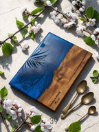 A rectangular wooden serving board with a blue resin section and a palm leaf design. The board is accompanied by three antique gold spoons and is decorated with white flowers and green leaves.