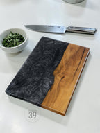 A rectangular cutting board with a black and wood grain design. A stainless steel knife and a small bowl of green herbs are also present. The cutting board has the text "ART STUDIO 39" printed on it.