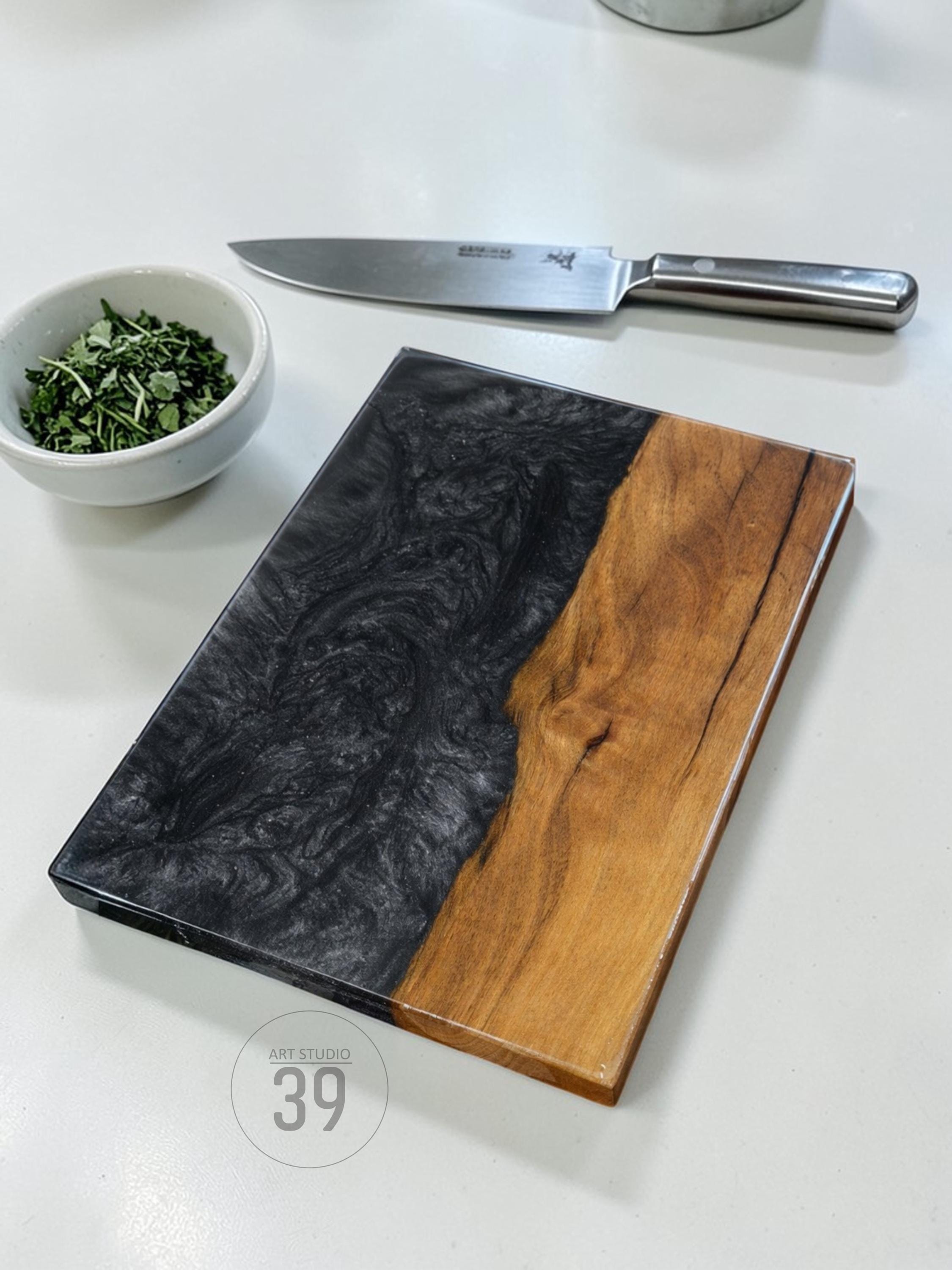 A rectangular cutting board with a black and wood grain design. A stainless steel knife and a small bowl of green herbs are also present. The cutting board has the text "ART STUDIO 39" printed on it.