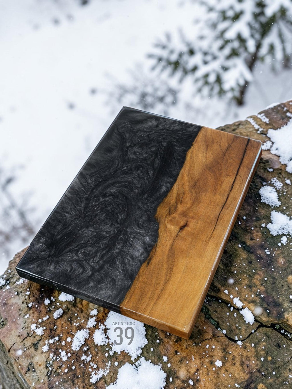 Rectangular wooden serving board with a striking design. One half features a swirling black epoxy resin, while the other showcases natural wood grain. The board is set against a snowy backdrop, with the text "ART STUDIO 39" visible.