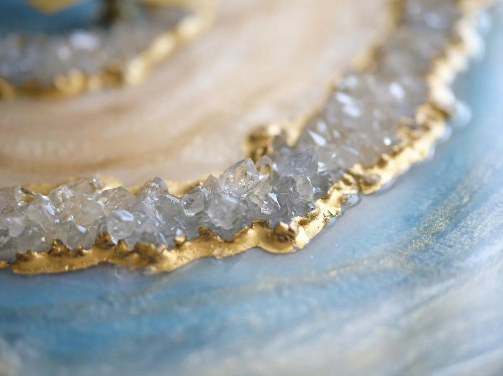 Close-up of a decorative resin piece with a gold-painted edge and embedded crystals. The piece has a light blue and cream color scheme, with a shimmering, polished finish. The crystals are clear and add texture.
