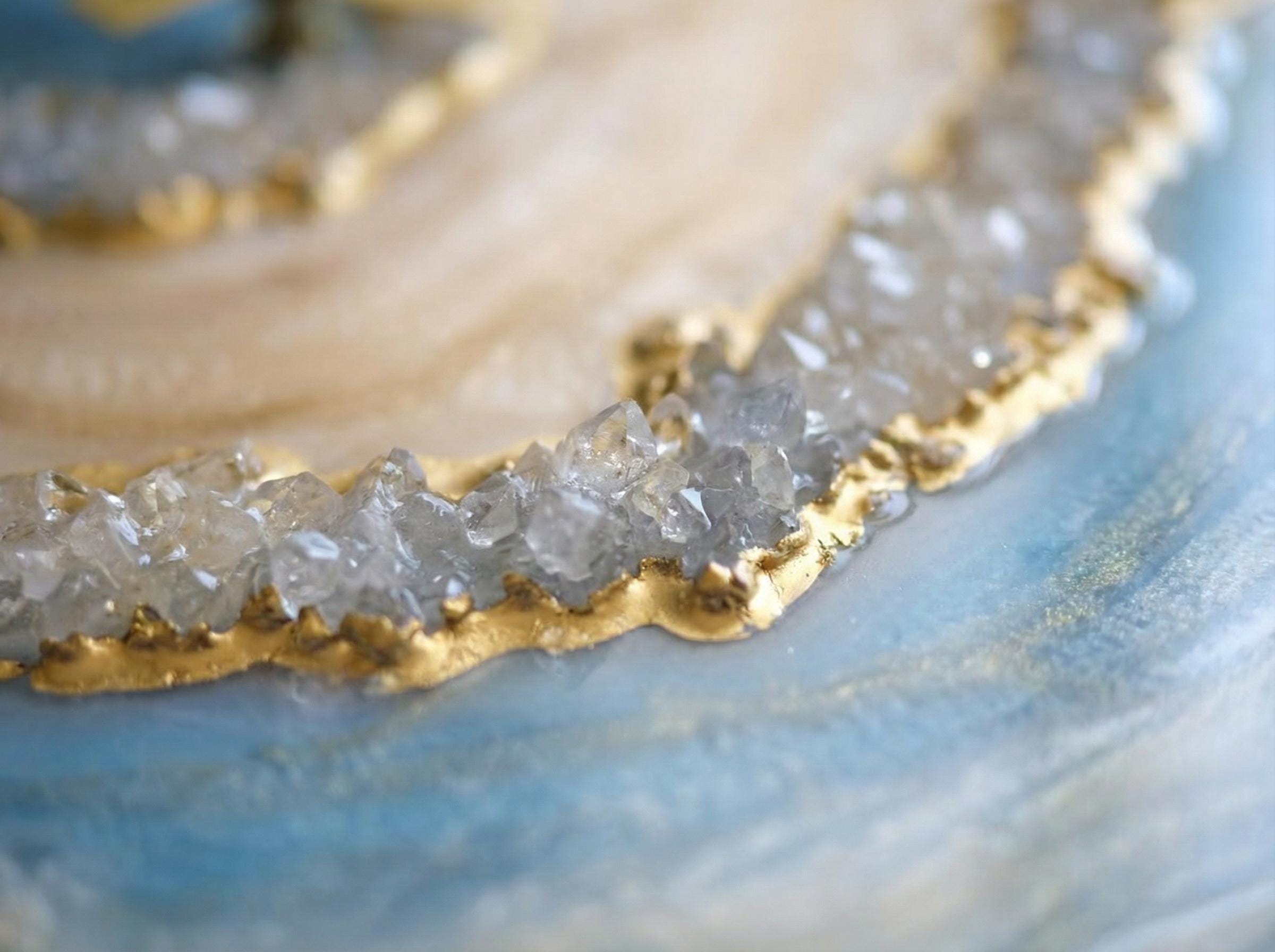 Close-up of a decorative resin piece with a gold-painted edge and embedded crystals. The piece has a light blue and cream color scheme, with a shimmering, polished finish. The crystals are clear and add texture.