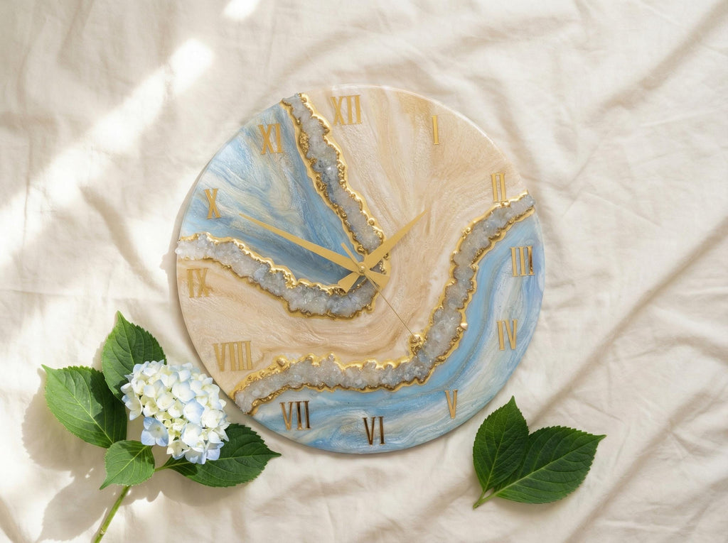 Round wall clock with a blue, beige, and gold resin design. The clock face features Roman numerals and gold clock hands. The clock is decorated with a blue and white hydrangea flower and green leaves.