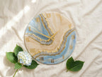 Round wall clock with a blue, beige, and gold resin design. The clock face features Roman numerals and gold clock hands. The clock is decorated with a blue and white hydrangea flower and green leaves.