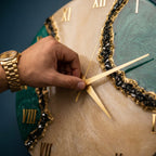 A round wall clock featuring a beige marbled face with deep green marbled sections, accented by textured black crystalline borders. The clock has gold-colored Roman numerals and thin gold-colored hands, with a gold-tone metal watch.