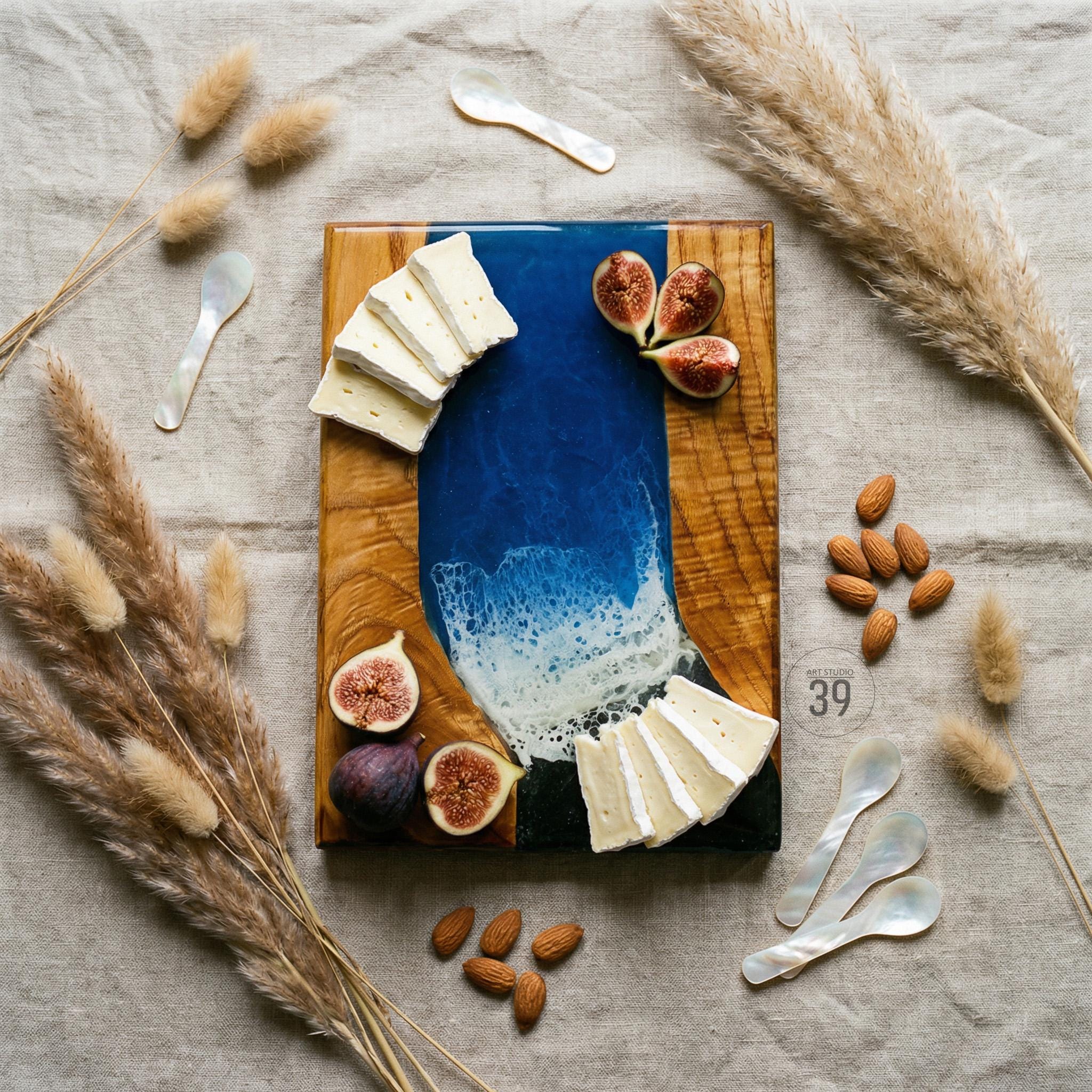 A wooden serving board with a blue resin wave design, adorned with brie cheese, figs, and almonds. The board is accompanied by mother-of-pearl spoons and decorative wheat stalks, creating an elegant presentation.