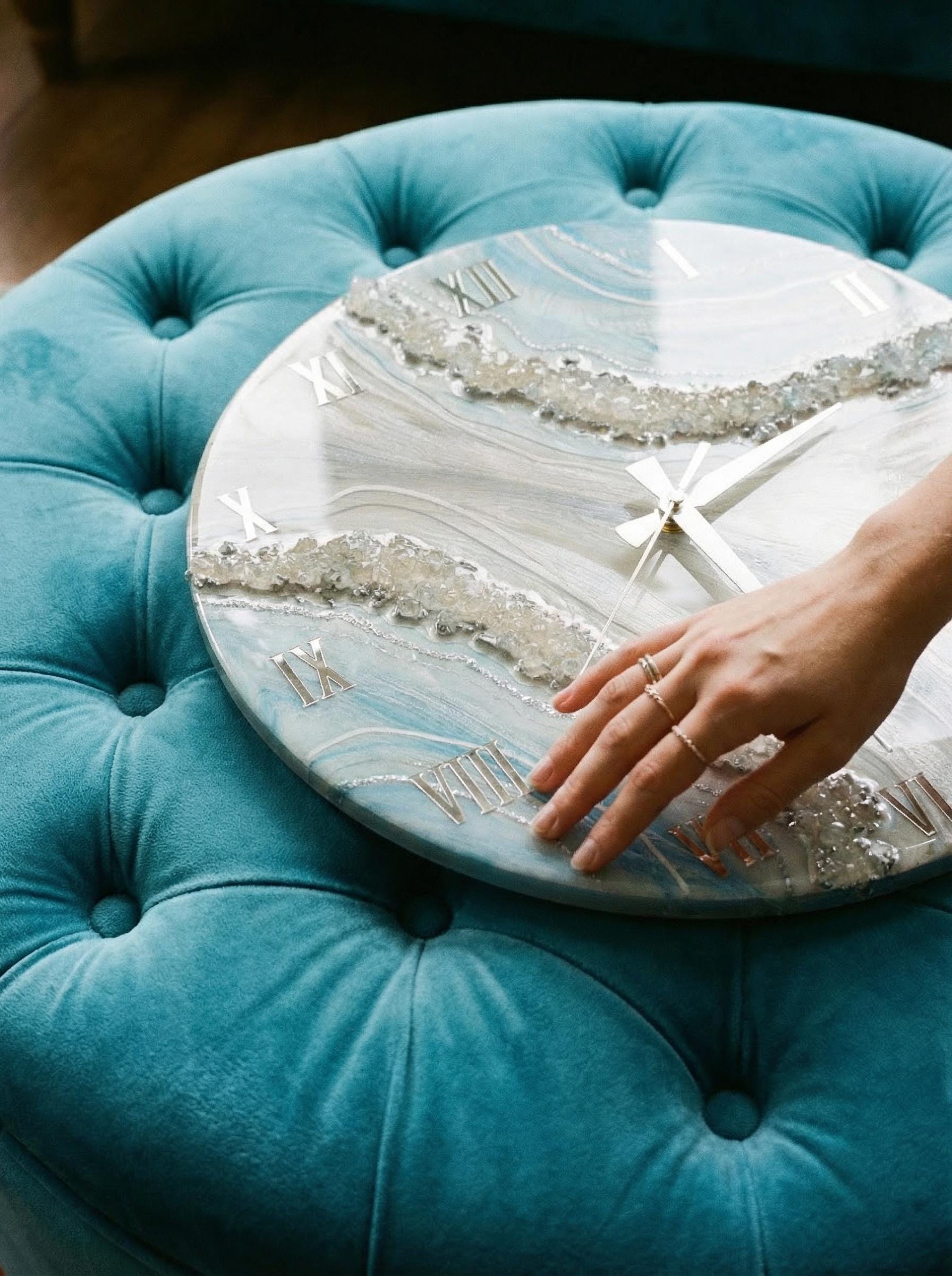 A round wall clock with a blue and white marbled design, silver Roman numerals, and silver clock hands. The clock has a decorative crystal-like border. The clock is resting on a teal upholstered ottoman.
