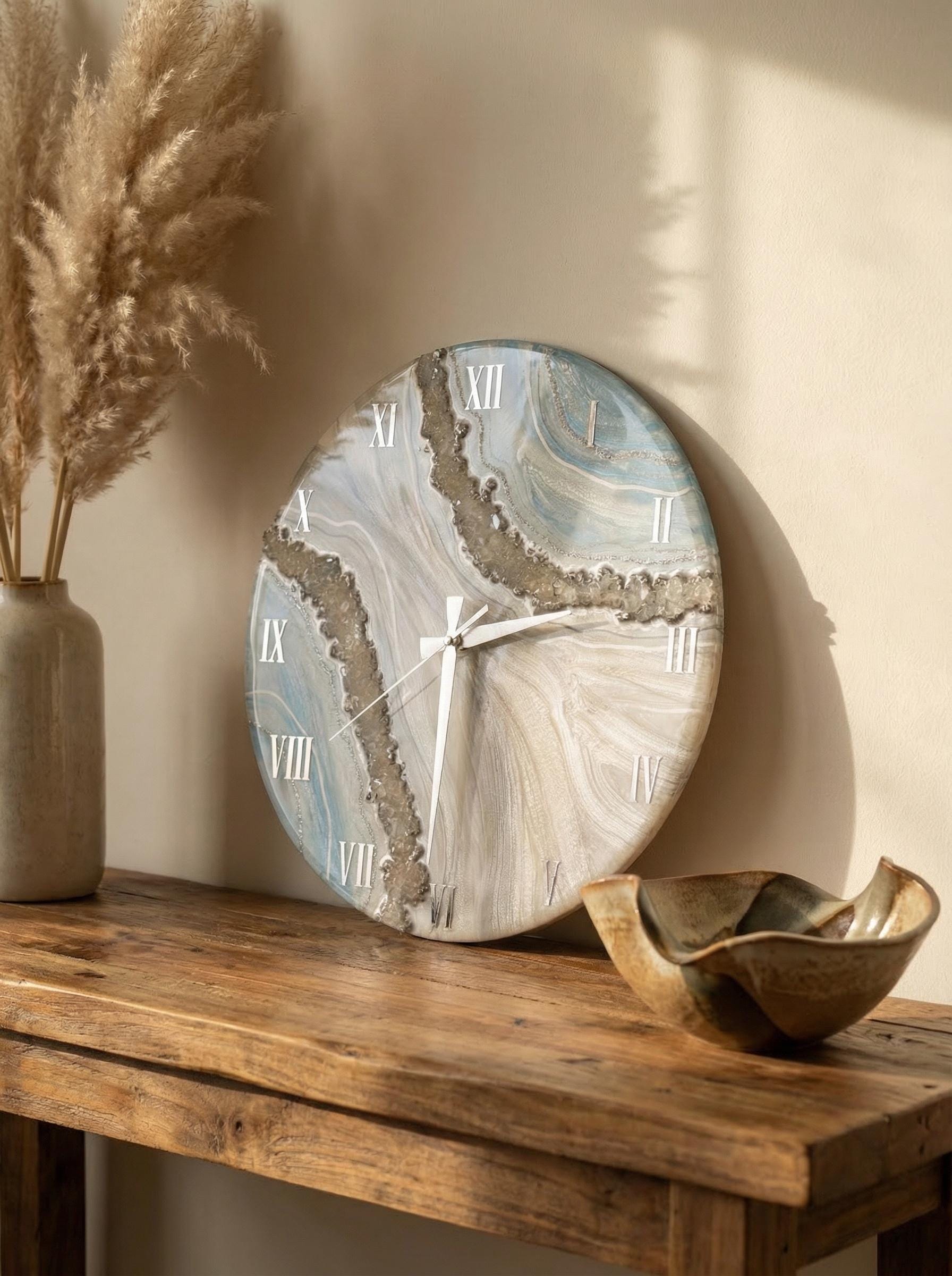 A round wall clock with a marbled design in shades of blue, white, and gray. The clock features white Roman numerals and hands. The clock is displayed on a wooden table with a decorative vase and bowl.