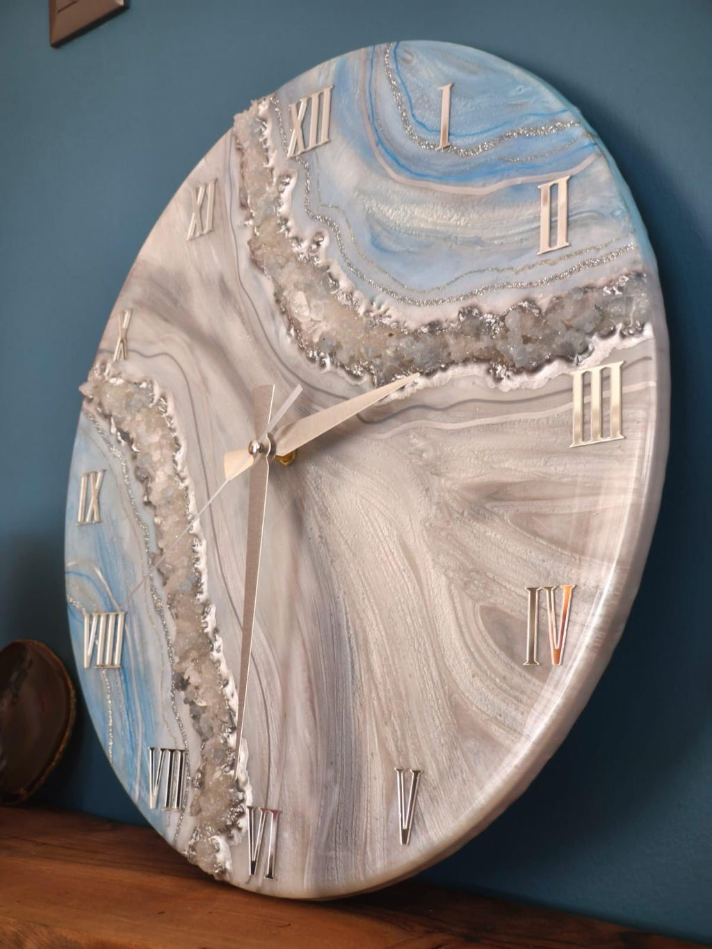 Round wall clock with a marbled design in shades of blue, gray, and white. The clock features silver Roman numerals and hands. The clock's design includes a sparkling, crystal-like border.