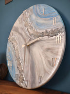 Round wall clock with a marbled design in shades of blue, gray, and white. The clock features silver Roman numerals and hands. The clock's design includes a sparkling, crystal-like border.