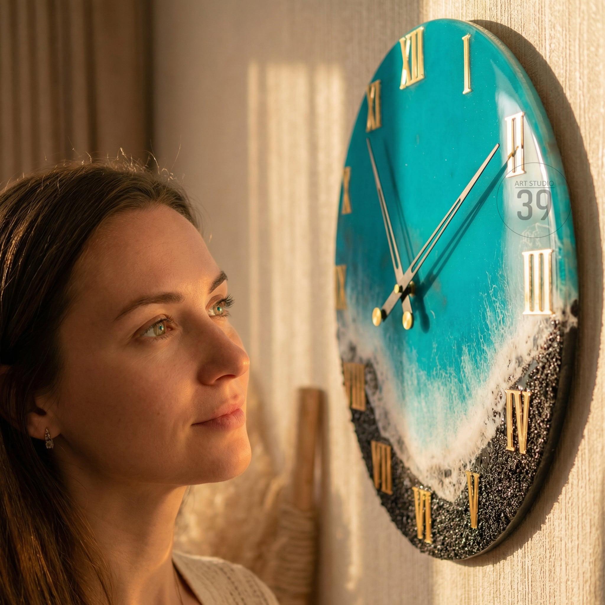 Round wall clock with a turquoise face, gold Roman numerals, and gold hands. The clock face features a wave design with white foam and black sand. The text "ART STUDIO 39" is visible on the clock face.
