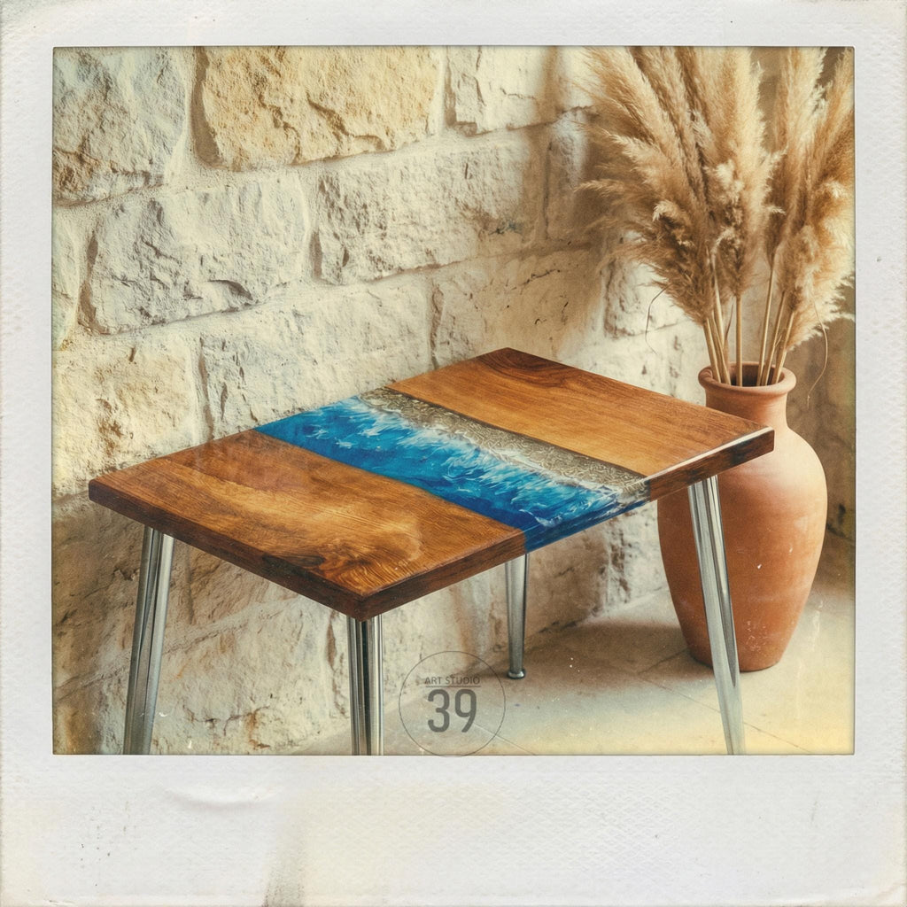 A handcrafted wooden side table with a unique ocean-inspired design. The tabletop features sections of natural wood and a blue resin section resembling waves. The table has chrome legs and is placed near a stone wall and a terracotta vase.