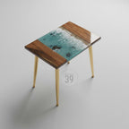 A small, rectangular side table with a unique design. The tabletop features a turquoise resin ocean scene with white waves, framed by dark wood. The table has gold-colored, tapered legs. The text "ART STUDIO 39" is in a circle on the table.