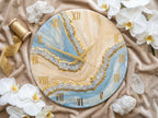 Round wall clock with a cream, blue, and gold resin design. The clock face features gold Roman numerals and hands. The design includes a geode-like pattern with crystal accents. Decorative elements include white orchids and crystals.