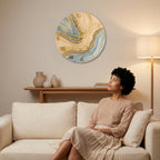 A round wall clock with a unique design. The clock face features a blue, beige, and gold abstract pattern. Gold Roman numerals mark the hours. The clock hands are gold. The clock is hanging on a light-colored wall.