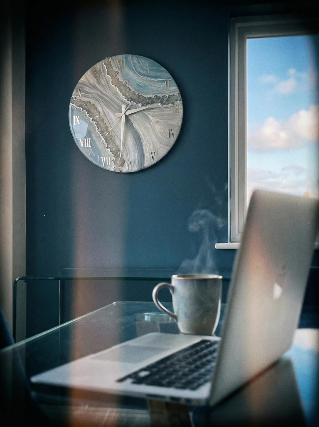 A round wall clock with a blue, gray, and white marbled design hangs on a dark blue wall. The clock has white Roman numerals. A laptop and a mug of coffee sit on a glass table.
