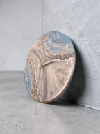 An oval-shaped wall clock with a marbled design in shades of blue, gray, and white. The clock face features Roman numerals in a metallic gold color and matching clock hands. The clock is leaning against a gray concrete wall.