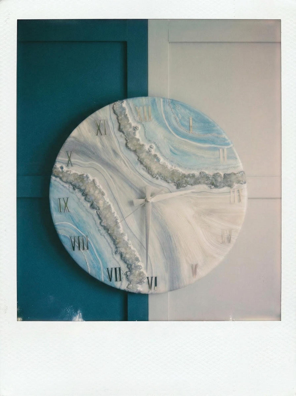Round wall clock with a blue, white, and gray marbled design. The clock face features Roman numerals and silver-colored hands. The clock has a decorative, textured border and is mounted on a wall.