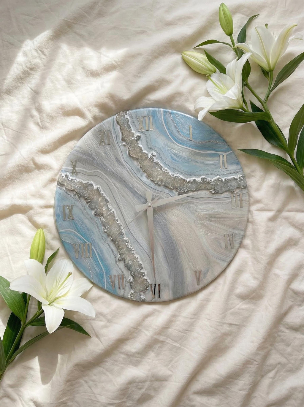 Round wall clock with a blue, gray, and white marbled design. The clock features silver Roman numerals and a silver border. The clock hands are silver. The clock is surrounded by white lilies.