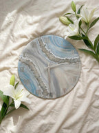Round wall clock with a blue, gray, and white marbled design. The clock features silver Roman numerals and a silver border. The clock hands are silver. The clock is surrounded by white lilies.