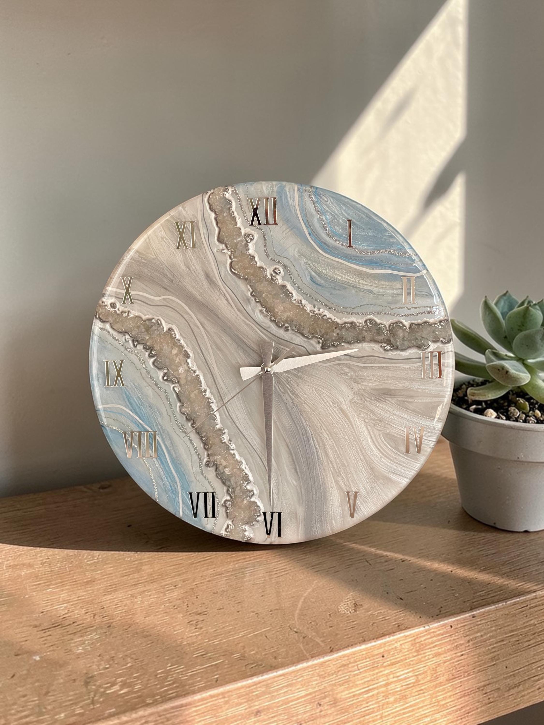 Round wall clock with a marbled design in shades of blue, gray, and white. The clock features Roman numerals in a metallic gold color and silver clock hands. A succulent plant in a gray pot sits to the right.