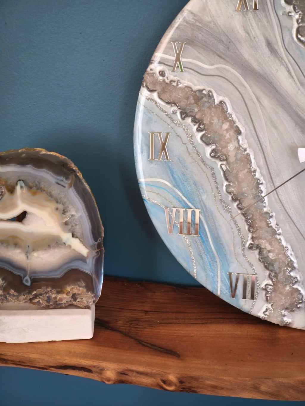 A decorative wall clock with a marbled design in shades of gray and blue, featuring Roman numeral hour markers. The clock is displayed with a natural agate stone on a wooden shelf.
