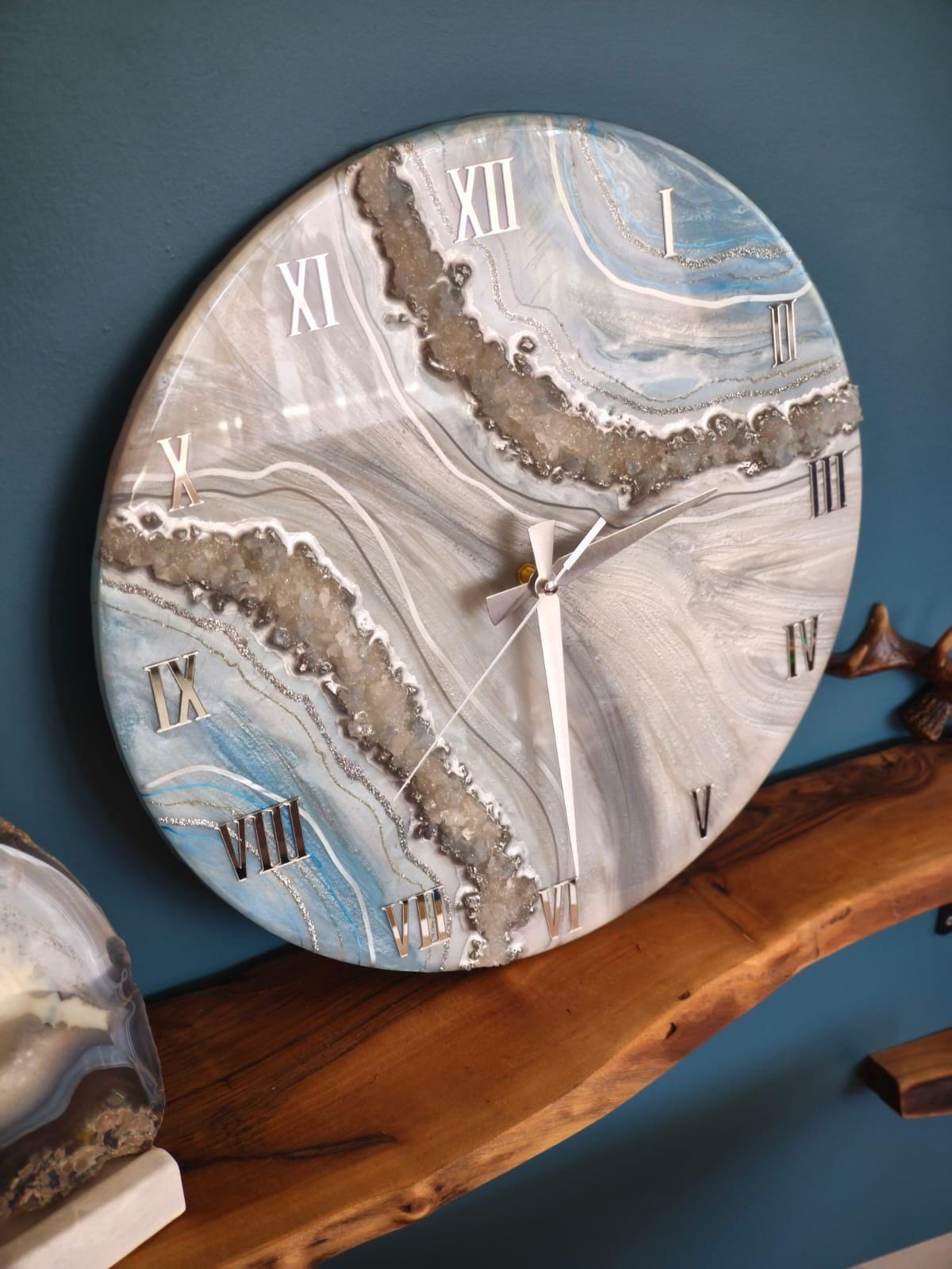Round wall clock with a marbled design in shades of blue, gray, and white. The clock face features silver Roman numerals and hands. A decorative, modern timepiece for home decor.